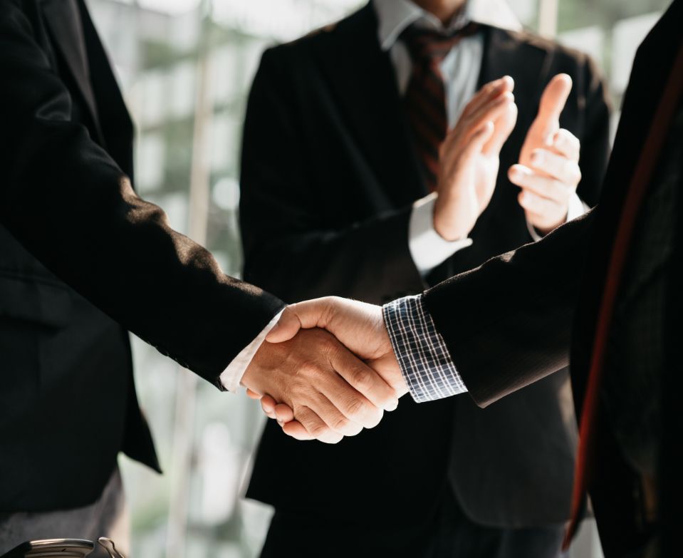 Business men shaking hands with another man in background applauding.